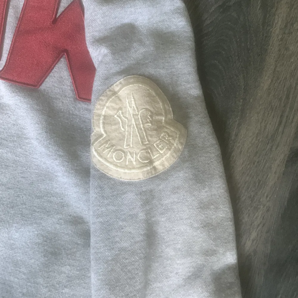 Säljer en snygg grå sweatshirt från Moncler med texten 'NOW' i rött på framsidan. Tröjan har långa ärmar och en klassisk rund halsringning. Perfekt för en casual look eller att mysa i hemma. Moncler-loggan finns på ärmen för en extra touch av stil. . Tröjor & Koftor.