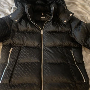 Svart pufferjacka från Boss - Snygg svart pufferjacka från Boss med diskret mönster och dragkedja framtill. Jackan har en huva och praktiska fickor med dragkedjor. Perfekt för kyligare dagar. Kvitto Medföljs av Köp, STRL L