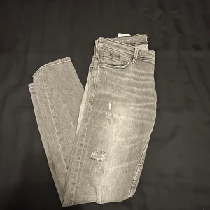 Grå jeans med slitningar - Snygga grå jeans från Icono Couture med coola slitningar för en trendig look. De har en klassisk femficksdesign och en knappgylf. Perfekta för en avslappnad stil.