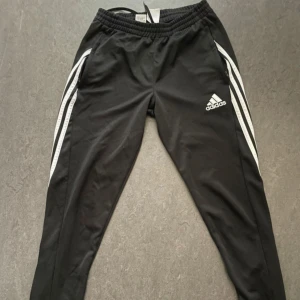 Svarta träningsbyxor från Adidas - Snygga svarta träningsbyxor från Adidas med vita ränder längs sidorna. Byxorna har en elastisk midja med snörning och praktiska dragkedjor vid bensluten. Perfekta för träning eller en avslappnad dag. Finns ett litet håll inåt, som inte alls syns - visas på sista bilden 