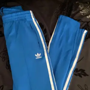 Snygga blå träningsbyxor från Adidas med vita ränder längs sidorna. Byxorna har en elastisk midja och dragkedja vid fickorna för extra funktionalitet. Perfekta för en sportig look eller träning. Använt väldigt få gånger 