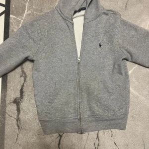 Grå huvtröja från Ralph Lauren - Säljer en grå huvtröja från Ralph Lauren med dragkedja och broderad logga på bröstet. Tröjan har långa ärmar och en bekväm passform. Perfekt för en avslappnad stil.