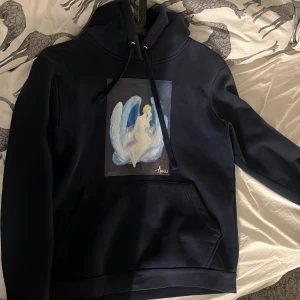 Svart hoodie med ängelmotiv från The Cool elephant Elephant - Säljer en svart hoodie med ett unikt ängelmotiv på framsidan från The Cool Elephant. Tröjan har en klassisk design med dragsko i huvan och en stor ficka framtill. Perfekt för en stilren och bekväm look.