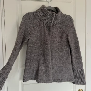 Grå stickad tröja från Lindex - Säljer en mysig grå stickad tröja från Lindex med hög krage och knappar framtill. Tröjan har långa ärmar och ribbade detaljer vid krage och ärmslut. 