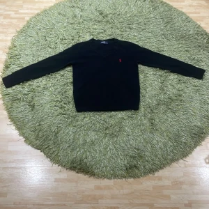 Svart tröja från Polo Ralph Lauren - Säljer en stilren svart tröja från Polo Ralph Lauren med röd broderad logga på bröstet. Tröjan har en v-ringad design och långa ärmar, perfekt för en klassisk look.