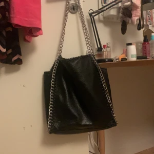 Svart axelväska med kedjedetaljer - Jätte fin axelväska med kedjor, perfekt till skolan då den får plats med dator osv. Den är från tiamo och köpt på Sellpy, den har några defekter på baksidan (se bilder) där det har ”skavts upp” och en ”tråd” har lossnat, men den går att sy ihop om man vill. Defektena är inget man tänker på enligt mig och jag säljer då jag har en annan väska❤️❤️ kom privat vid frågor osv
