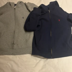 Grå och marinblå hoodies från Ralph Lauren - Två snygga hoodies från Ralph Lauren i storlek L för barn. En i klassisk grå och en i marinblå, båda med dragkedja och den ikoniska loggan på bröstet. Perfekta för en avslappnad stil. Skick 9,5/10 knappt använda. 1000 för båda