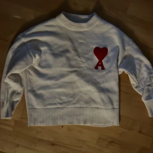 Vit tröja från Ami - Säljer en stilren vit tröja från Ami med ett rött hjärta och bokstaven 'A' på bröstet. Tröjan har långa ärmar och ribbade muddar vid ärmslut och nederkant. Perfekt för en avslappnad och trendig look.
