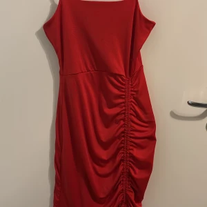 Röd klänning med rynkdetalj - Snygg röd klänning med rynkdetalj på sidan och smala axelband. Perfekt för en kväll ute eller en speciell tillställning. Klänningen har en figurnära passform och en liten volangkant längst ner.