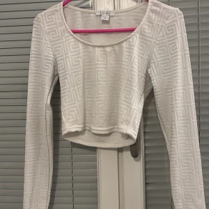 Vit långärmad topp från new yorker - Säljer en stilren vit långärmad topp från new yorker med ett diskret geometriskt mönster. Toppen är croppad och har en rund halsringning. Perfekt för att lägga till en touch av elegans till din outfit.