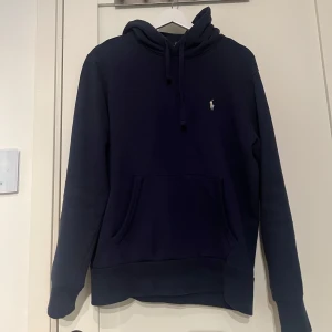 Mörkblå hoodie från Ralph Lauren - Säljer en stilren mörkblå hoodie från Ralph Lauren med det klassiska logotypbroderiet på bröstet. Tröjan har en känguruficka och justerbar huva med dragsko. Perfekt för en avslappnad stil. Köpt i november på farfetch för 2100kr. Kvitto och tags kommer med!