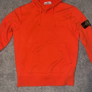 Orange hoodie från Stone Island - Säljer en snygg orange hoodie från Stone Island med klassisk logga på ärmen. Tröjan har en stor ficka framtill och justerbar huva med dragsko. Perfekt för en casual look. 🧡