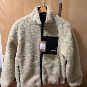 Stussy fleece jacka - Säljer en snygg beige teddyjacka från Stussy med svart ficka och dragkedja fram. Jackan har en cool 8-ball design på ryggen och är vändbar med en svart insida. Perfekt för att hålla sig varm och stilren.