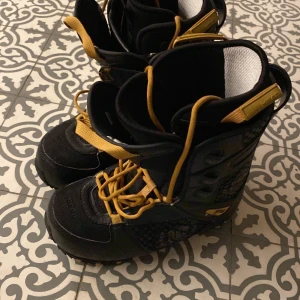 Svarta snowboard boots med gula detaljer - Säljer ett par svarta snowboard boots med gula snören och detaljer. De har en robust design med snörning och är perfekta för vintersport.