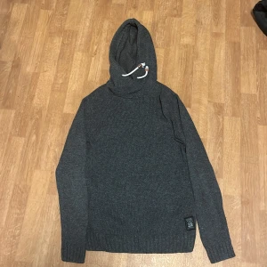 Grå stickad hoodie från John Devin - Säljer en stilren grå stickad hoodie från John Devin. Perfekt för kyliga dagar med sin mjuka och bekväma passform. Den har en justerbar huva med snören och en snygg detalj med Work Wear-märke på framsidan. Passar utmärkt till både vardag och avslappnade tillfällen.