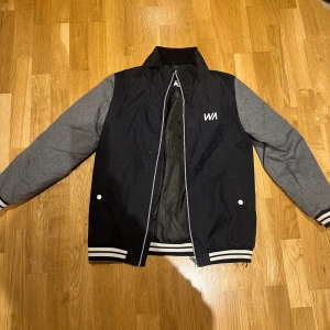 Svart och grå bomberjacka från Warp - Säljer en snygg svart och grå bomberjacka från Warp. Jackan har långa grå ärmar och en svart kropp med vita detaljer. Den har dragkedja framtill och två fickor med knappar. Perfekt för en cool och avslappnad stil.