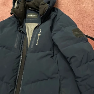 Mörkblå dunjacka från Sail Racing - Säljer en mörkblå dunjacka från Sail Racing med huva och dragkedja. Jackan har en stilren design med logotyp på bröstet och en patch på ärmen. Perfekt för kalla dagar.