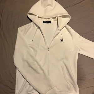 Vit hoodie från Ralph Lauren - Säljer en stilren vit hoodie från Ralph Lauren med dragkedja och den klassiska loggan på bröstet. Tröjan har en justerbar huva med snören och en praktisk ficka framtill. Perfekt för en avslappnad look.