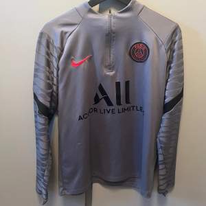 Snygg grå långärmad PSG-tröja från Nike med klubbens emblem på bröstet och sponsorlogga 'Accor Live Limitless'. Tröjan har en halv dragkedja framtill och stilrena mönster på ärmarna. Skriv om ni har frågor den har ett jätte litet hål på armen men inget som syns mycket 