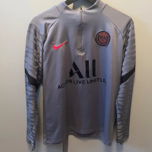 Grå långärmad PSG-tröja från Nike - Snygg grå långärmad PSG-tröja från Nike med klubbens emblem på bröstet och sponsorlogga 'Accor Live Limitless'. Tröjan har en halv dragkedja framtill och stilrena mönster på ärmarna. Skriv om ni har frågor den har ett jätte litet hål på armen men inget som syns mycket 