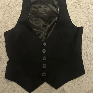 Svart väst med knappar - Snygg svart väst med sex knappar framtill. Perfekt för att ge en stilren touch till din outfit. Västen har en klassisk design och är lätt att matcha med olika plagg.