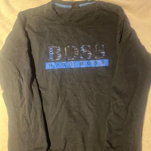 Svart långärmad tröja från Hugo Boss - Säljer en svart långärmad tröja från Hugo Boss med ett stilrent tryck i blått och svart på framsidan. Tröjan är lite sliten i kragen samt en aning solblekt (vilket syns på bilderna) 
