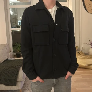 Svart overshirt från Zara - Säljer en stilren svart overshirt från Zara med knappar framtill och två stora bröstfickor. Perfekt för lager-på-lager-stil och passar till många olika outfits. Enkelt och snyggt plagg för alla tillfällen.