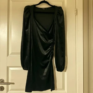 Svart omlottklänning från Bik Bok - Säljer en elegant svart omlottklänning från Bik Bok i storlek XS. Klänningen har långa ärmar och en smickrande omlottdesign som ger en fin siluett. Perfekt för festliga tillfällen eller en utekväll. Materialet har en lätt glansig finish som ger en lyxig känsla.