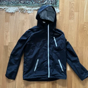 Svart vindjacka från Craft - Säljer en svart vindjacka från Craft. Den har en praktisk huva och dragkedja framtill. Jackan är perfekt för blåsiga dagar och har en stilren design med en bröstficka med dragkedja. Passar bra för utomhusaktiviteter och är i bra skick.