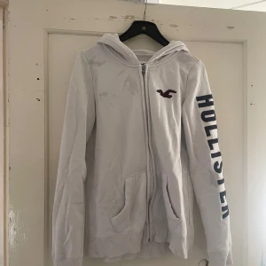 Vit hoodie från Hollister - Säljer en vit hoodie från Hollister i storlek S. Den har en dragkedja framtill och en stor logga på ärmen. Perfekt för en avslappnad stil och passar bra till både jeans och mjukisbyxor. Den är i bra skick och supermjuk! 🕊️