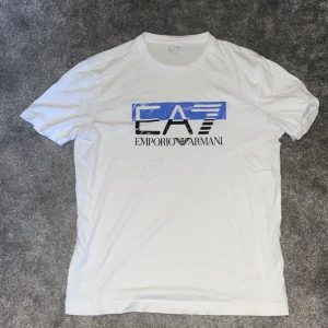 Vit t-shirt från Emporio Armani - Säljer en snygg vit t-shirt från Emporio Armani med deras ikoniska logga i blått och svart på framsidan. T-shirten är kortärmad och perfekt för en casual look. Passar bra till både jeans och shorts. Perfekt för sommaren! 🌞