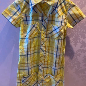 Rutig klänning från Encore - Säljer en superfin gul och blå rutig klänning från Encore i storlek XS.✨ Skjortan har korta ärmar och två bröstfickor, tyvärr utan bandet till. Den är i nyskick och redo för nya äventyr! 🌞💫