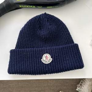 Säljer en snygg mörkblå ribbad mössa från Moncler. Den är perfekt för kyliga dagar och har en stilren design med Monclers logga framtill. Mössan är i nyskick och passar perfekt till både vardag och vinteraktiviteter. Håll dig varm och trendig! 💙