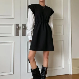 Svart kortklänning med vita ärmar - Snygg svart kortklänning med vita långa ärmar. Klänningen har en elastisk midja som ger en fin passform och en stilren look. Perfekt för både vardag och festliga tillfällen. Kombinera med stövlar för en komplett outfit!