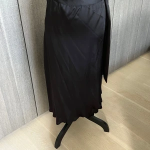 Svart omlottkjol - Snygg svart omlottkjol i ett elegant och flytande material. Perfekt för både vardag och festliga tillfällen. Kjolen har en stilren design med en smickrande passform som passar de flesta kroppstyper.