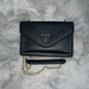 Svart axelväska från Guess - Snygg svart axelväska från Guess i skinn med guldkedja. Perfekt för både vardag och fest! Väskan har flera fack och stängs med en tryckknapp. Den är stilren och lätt att matcha med olika outfits.