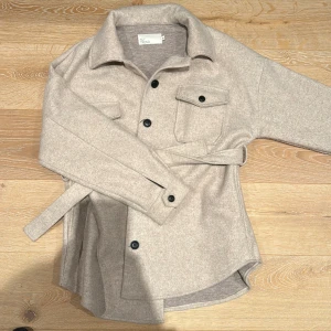 Beige overshirt / kappa från NLY Trend nelly  - Säljer en snygg beige overshirt från NLY Trend i storlek 36. Den har långa ärmar och knappar framtill samt ett bälte i midjan för en stilren look. Perfekt för höst och vår!