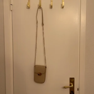 Beige axelväska med kedja - Säljer en superfin beige axelväska med en elegant kedja som axelrem. Väskan har en stilren design med en rund gulddetalj på framsidan. Perfekt för både vardag och fest! 👜✨