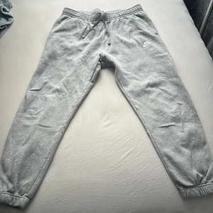 Grå mjukisbyxor från Nike - Säljer ett par grå mjukisbyxor från Nike i storlek M. De är superbekväma med elastisk midja och snörning för perfekt passform. Byxorna har en broderad Nike-logga på sidan och är gjorda av mjuk bomull, perfekt för chill dagar eller träning. 🩶
