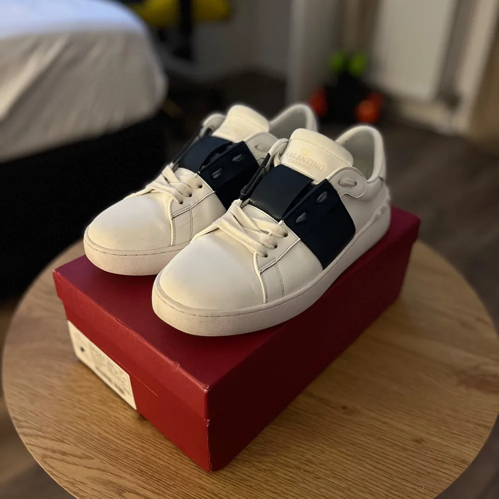 Säljer ett par snygga vita sneakers från Valentino med en cool svart detalj över snörningen. De är i nyskick och kommer i originalkartong. Perfekta för både vardag och fest! Storlek är inte specificerad, men de ser ut att vara i standardstorlek. Passa på att köpa dessa stilrena skor!. Skor.