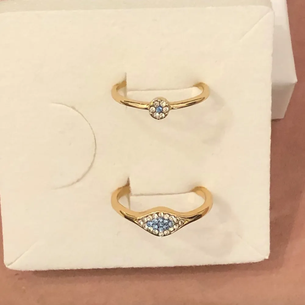 En ring med liten blomma i vitt och baby blå strass diamanter guld metall Och en ring med stenar i vitt och blått Dem passar fint tillsammans Inte använda pga fel storlek nickelfria . Asusteet.