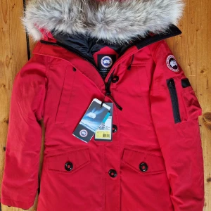  Canada Goose - Varm dunjacka från Canada Goose med löstagbar äkta päls på luvan. Fyra framfickor med skönt fleecefoder samt en ficka på vänster ärm och två praktiska innerfickor. Stängs med kraftig dragkedja och knappar. 