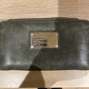 Marc by Marc Jacobs plånbok  - Säljer min fina plånbok som ej använts på ett tag. Silvrig bricka och dragkedja runtom. Många fack inuti, hel och ren. Lite skrapor på brickan som ej går att undgå. Härlig fin grågrön färg som är rätt unik.