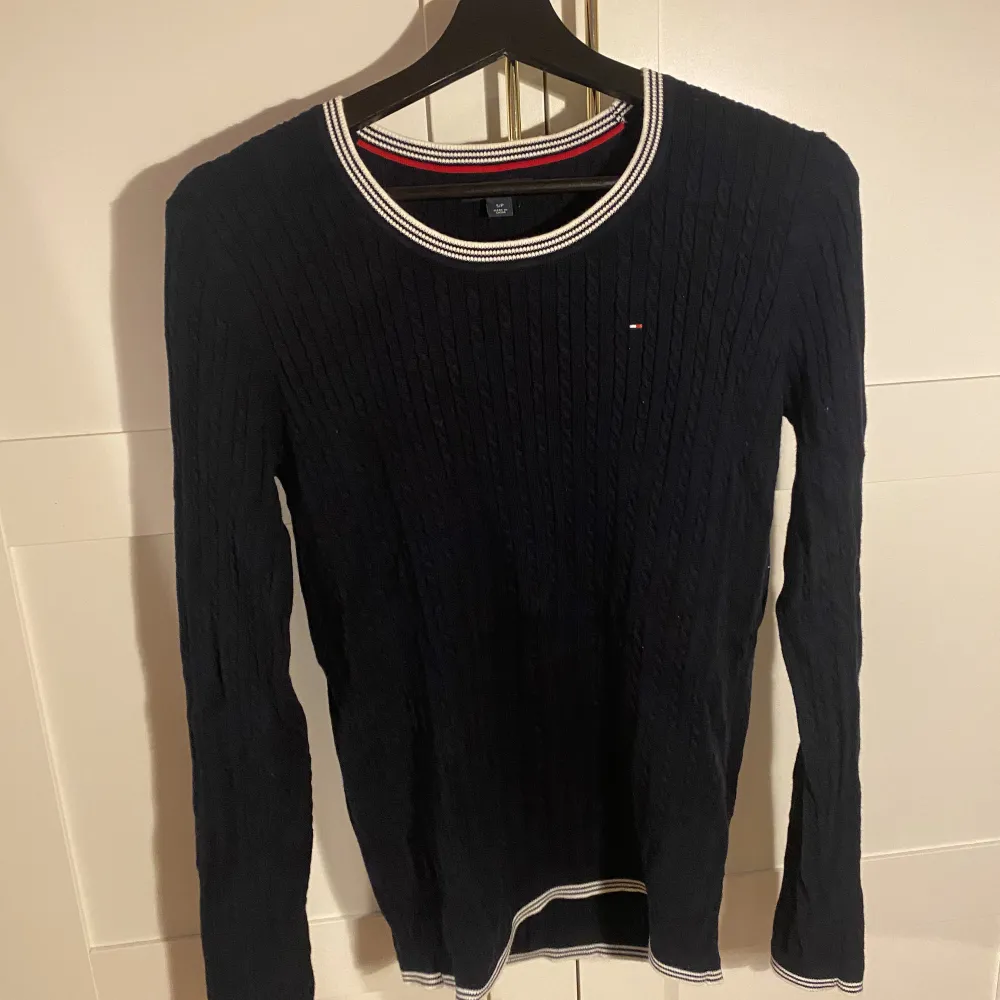 Marinblå kabelstickad tröja från Tommy Hilfiger, med vit/blå-randig detalj i krage och ärmar. Nypris 1199kr.. Neuleet.