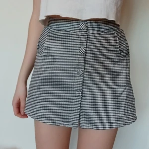 Minikjol med knappar - Svart/vit minikjol med knappar fram och fickor. Elastiskt midjebälte i bak och inbyggda safety shorts. Använd men i mycket bra skick