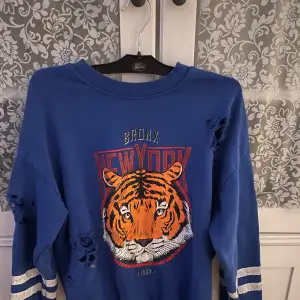 En blå sweatshirt från Ginatricot med ett tiger tryck på framsidan har tre hål som syns på bild 2&3 som fanns när jag köpte den 