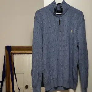 En kabelsticksd ljuslila/blå tröja med en zip up vid halsen. Gult märke på bröstet och en läder dragkedja! 
