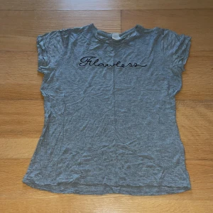 Grå GinaTricot topp - Kortärmad grå ginatricot topp med texten ”Flawless” i svart på framsidan. Har ett litet hål långt ner på höger sida. Priset inkluderar frakt (39kr). 