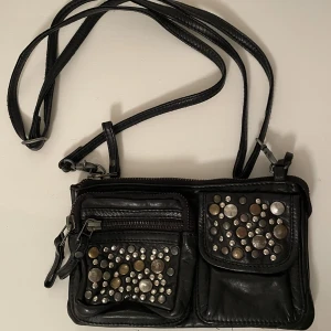 Fin axelremsväska - Mindre axelremsväska i läder med fina detaljer. Har reglerbart axelband så man kan välja längden/hur den ska sitta på själv. På insidan har den ett fack i mitten och och ett sidofack. Sparsamt använd, i väldigt fint skick.