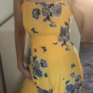 Gul/blå blommig maxiklänning  - Gul lång sommar klänning med blåa blommor och dekorativa knappar vid sidan  Storlek: 34 Använd 1 gång, nyskick. 